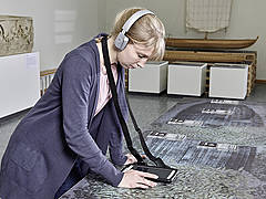 Besucherin berührt mit Handheld einen Infopoint und hört über Bluetooth-Kopfhöhrer weiterhführende Informationen zum Schiffswrack